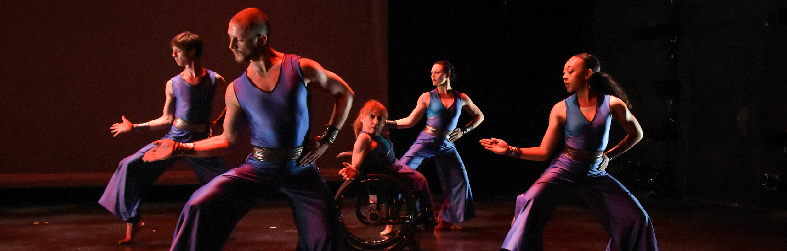 Two dancers, one standing, one in a wheelchair, crossing an arm with eachother, with a single dancer turning behind them