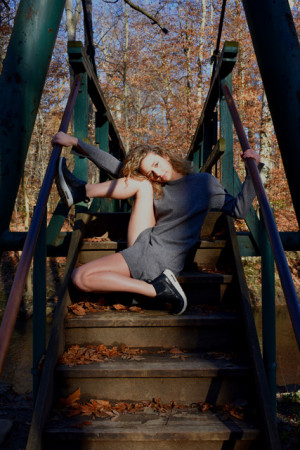 L. Taylor Ashton wears gray shorts jumpsuit and sneakers, posed outside on a set of steps with foot and hands on green handrails.