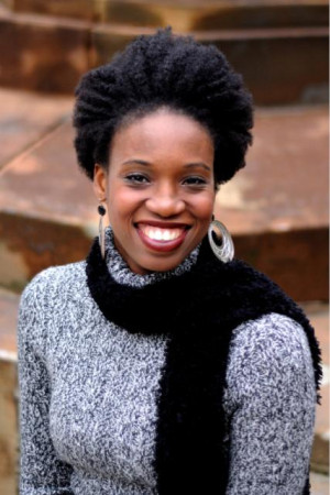 Andrea Belser McCormick wears a gray sweater, black scarf, and silver dangling earrings. She smiles wide, posed in front of red stone steps.