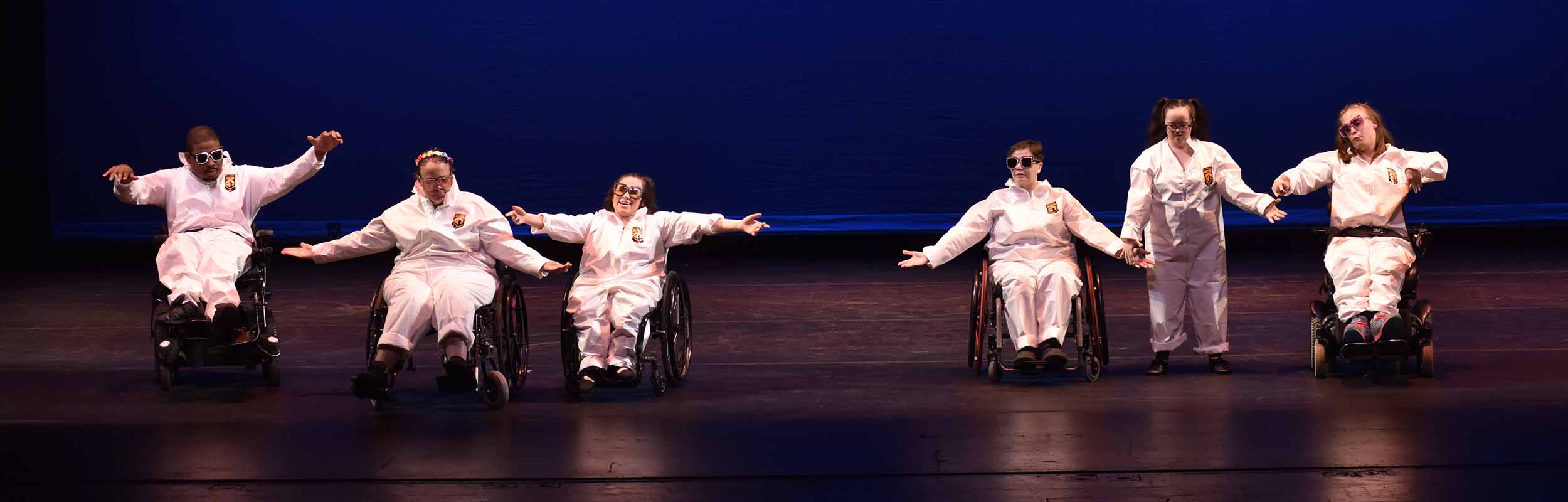 Dancers in the School of Dancing Wheels' Performance Ensemble performing in white astronaut-looking costumes and rainbow dazzled sunglasses