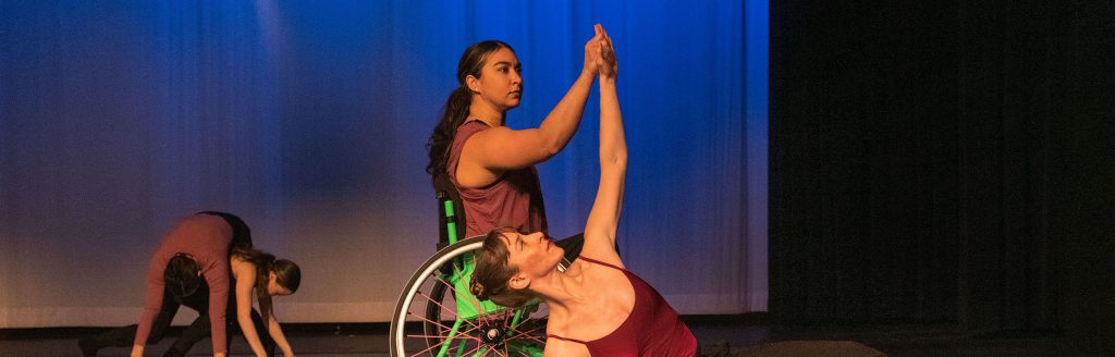 On a stage, company dancer Kimberly, who uses a wheelchair, holds the arm of sideways laying partner and company dancer Sara