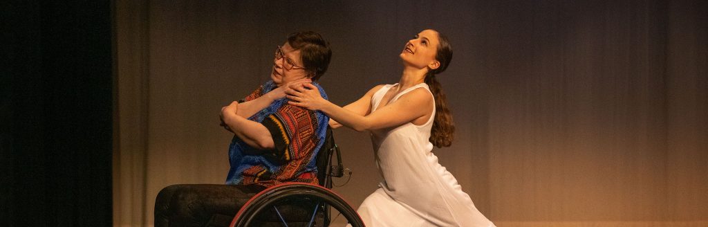 A dancer in a wheelchair embraced by a  standing dancer from behind
