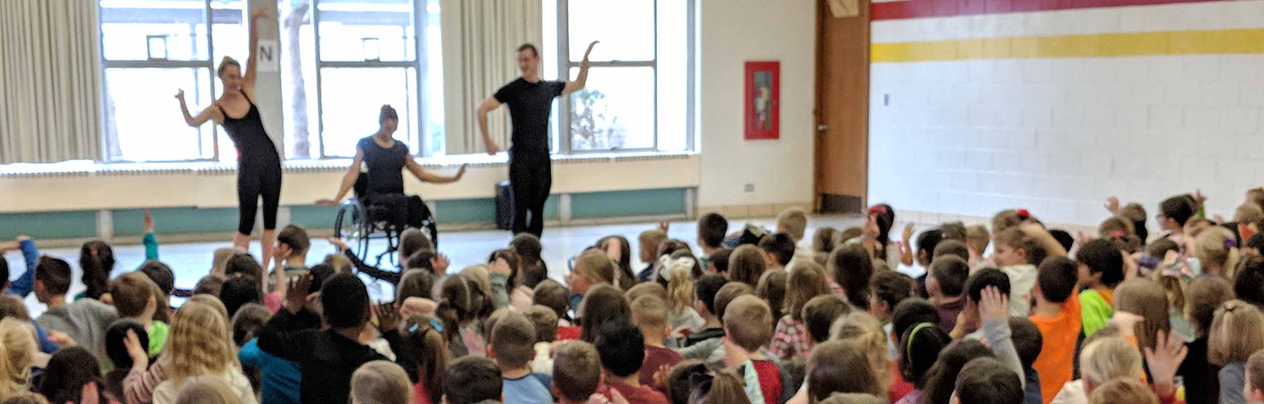Three company dancers teach arm movements to a large audience of elementary school students in an assembly style lecture performance.