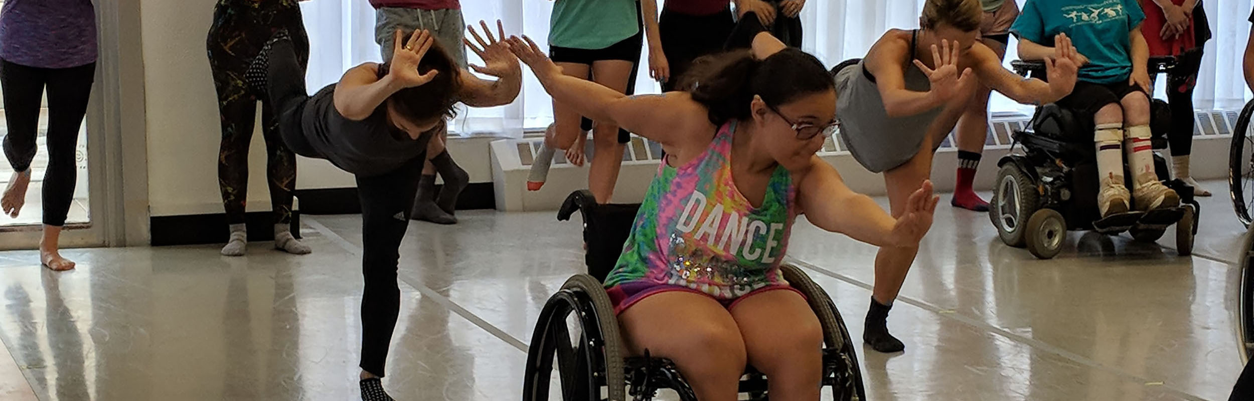 A sit-down dancer moves across the floor in a dance studio with stand-up dancers. Dancers of all abilities wait their turn behind them.