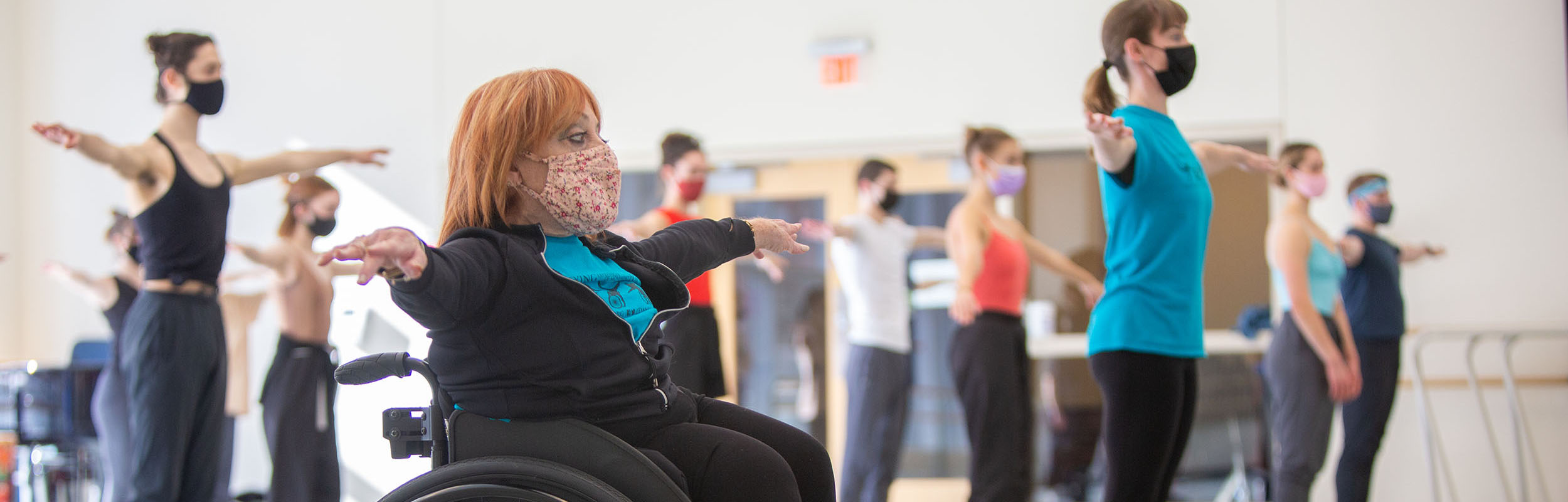 Physically integrated dance masterclass with Mary, a sit-down wheelchair dancer, and Sara, a stand-up dancer, teaching and students with arms out.