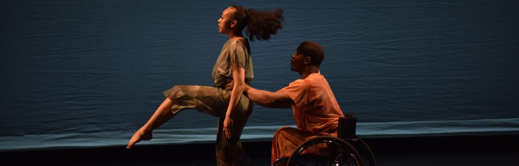 Performance photo of company dancer DeMarco in a wheelchair lifting partner Marissa in a bent leg shape from behind