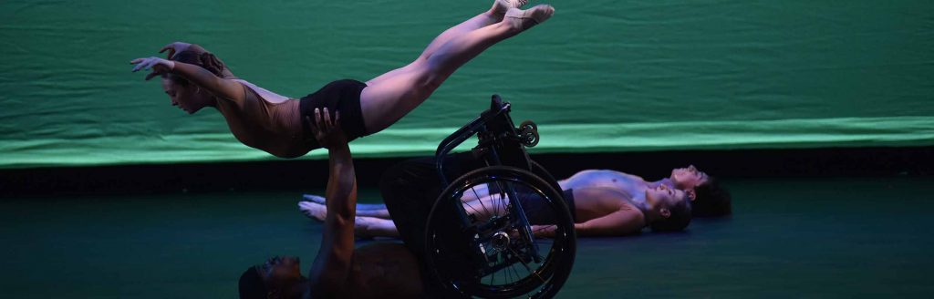 Company dancer DeMarco on his back in his wheelchair full-press lifting company dancer McKenzie in a flying position.