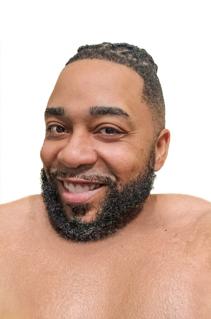 Craig Sams' headshot shows him with medium skin tone, bare shoulders, a gentle smile, facial hair over his lip and on his chin, and tightly braided hair shaved short on the sides of his head.