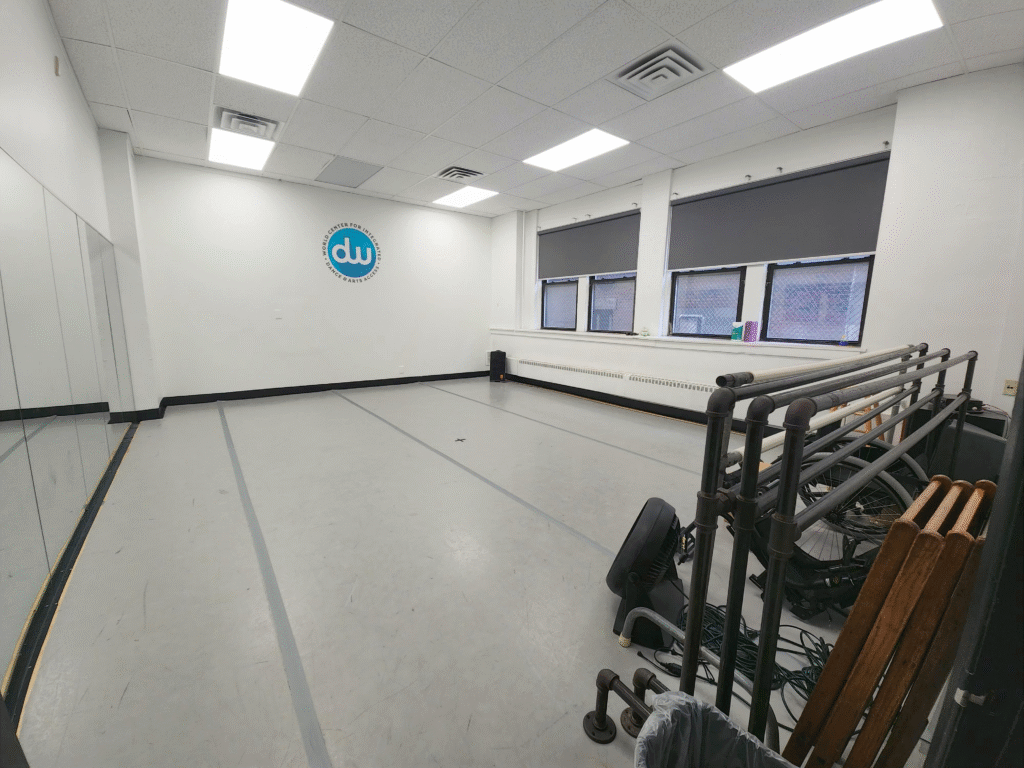 Image of a dance studio from the door in the lower left corner. There's a mirror on the wall to the left, a white wall across the room, and a wall with windows at the back. Bars and chairs are stored next to the wall closest to the door.